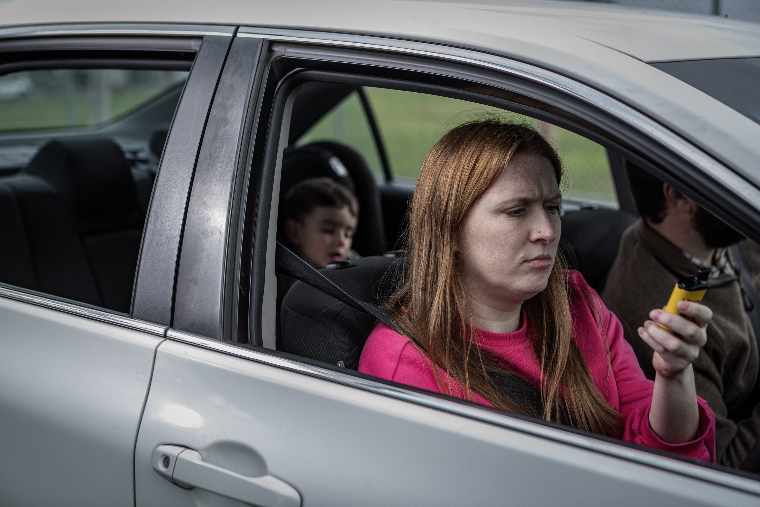 Young female driver of a car has a vape in her hand while a child is in the back seat.
